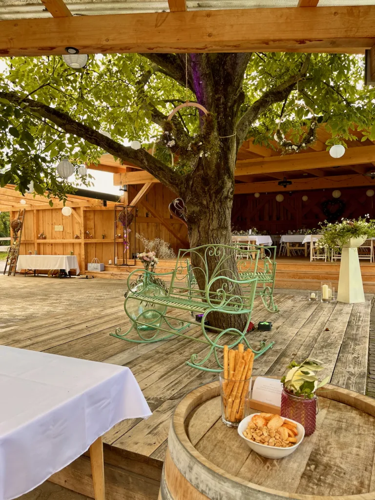 Bruders Schopf Oberkirch: Holzterrasse mit großem Baum, grüner Bank, Snacks auf Weinfass und festlich dekorierter Außenbereich.