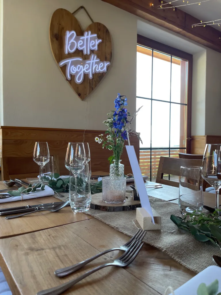 Hochzeit im Nillhof Fischerbach: Rustikaler Holztisch mit Blumendeko, Kerze, „Better Together“-Neonschild im Hintergrund.