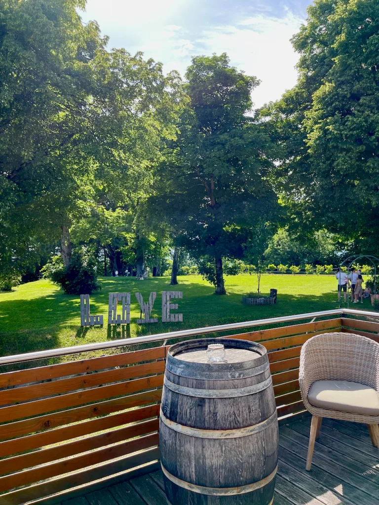 Außenbereich des Nillhofs Fischerbach: Große grüne Wiese mit LOVE-Buchstaben, Bäumen und rustikaler Terrasse mit Fass und Sitzgelegenheit.